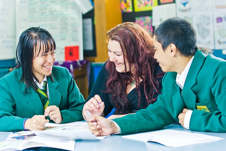 Female and male students in uniform at table with school work talking to female teacher